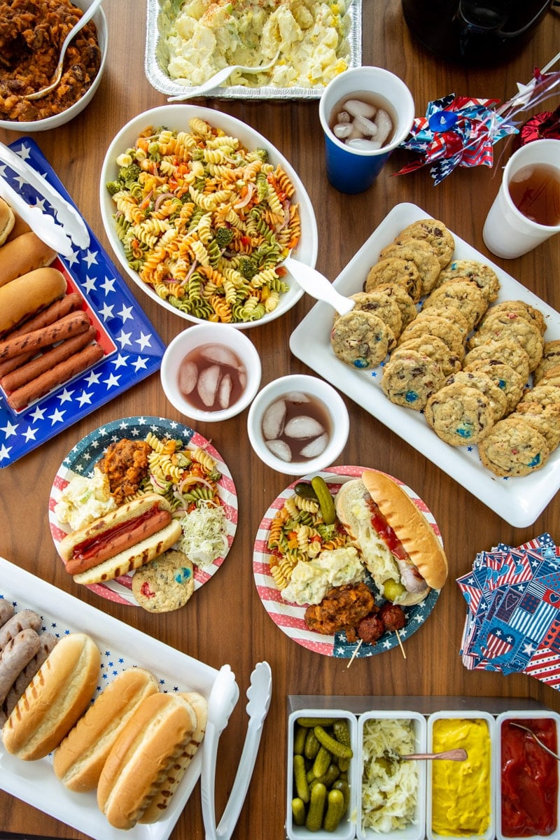 A table of 4th of July Cookout food such as brats, hot dogs, pasta salad, cookies, and drinks.