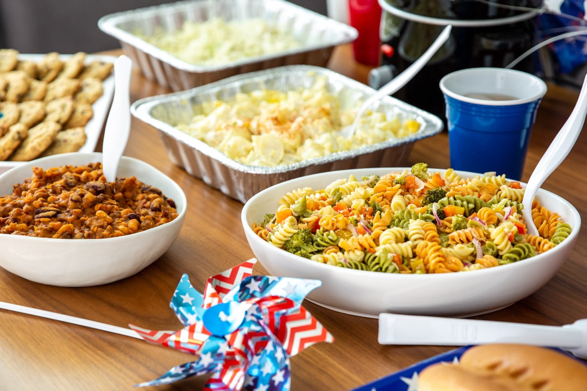 A table of 4th of July Cookout food such as brats, hot dogs, pasta salad, cookies, and drinks.