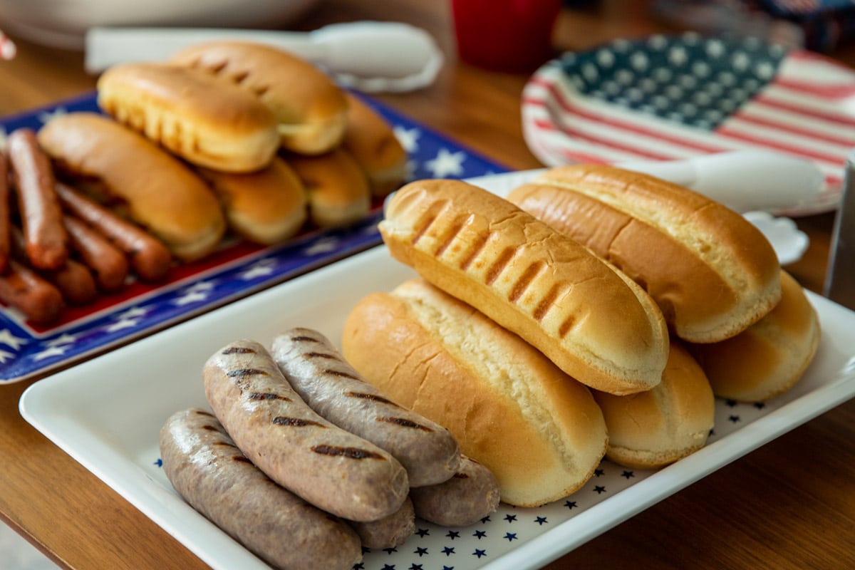 A table of 4th of July Cookout food such as brats, hot dogs, pasta salad, cookies, and drinks.