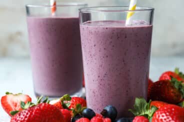 Two acai smoothies in clear glasses on a countertop surrounded by berries.