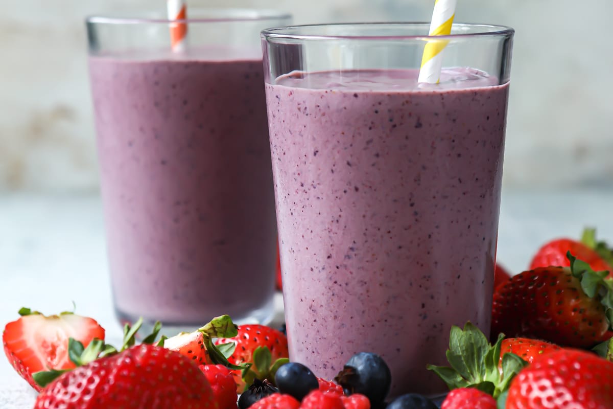 Two acai smoothies in clear glasses on a countertop surrounded by berries.