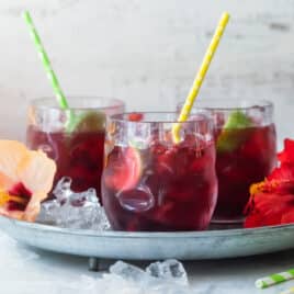 Glasses of Agua de Jamaica on a tray with hibiscus flowers and straws.