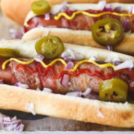 Bacon wrapped hot dogs on a wooden cutting board with toppings.