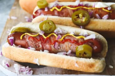 Bacon wrapped hot dogs on a wooden cutting board with toppings.