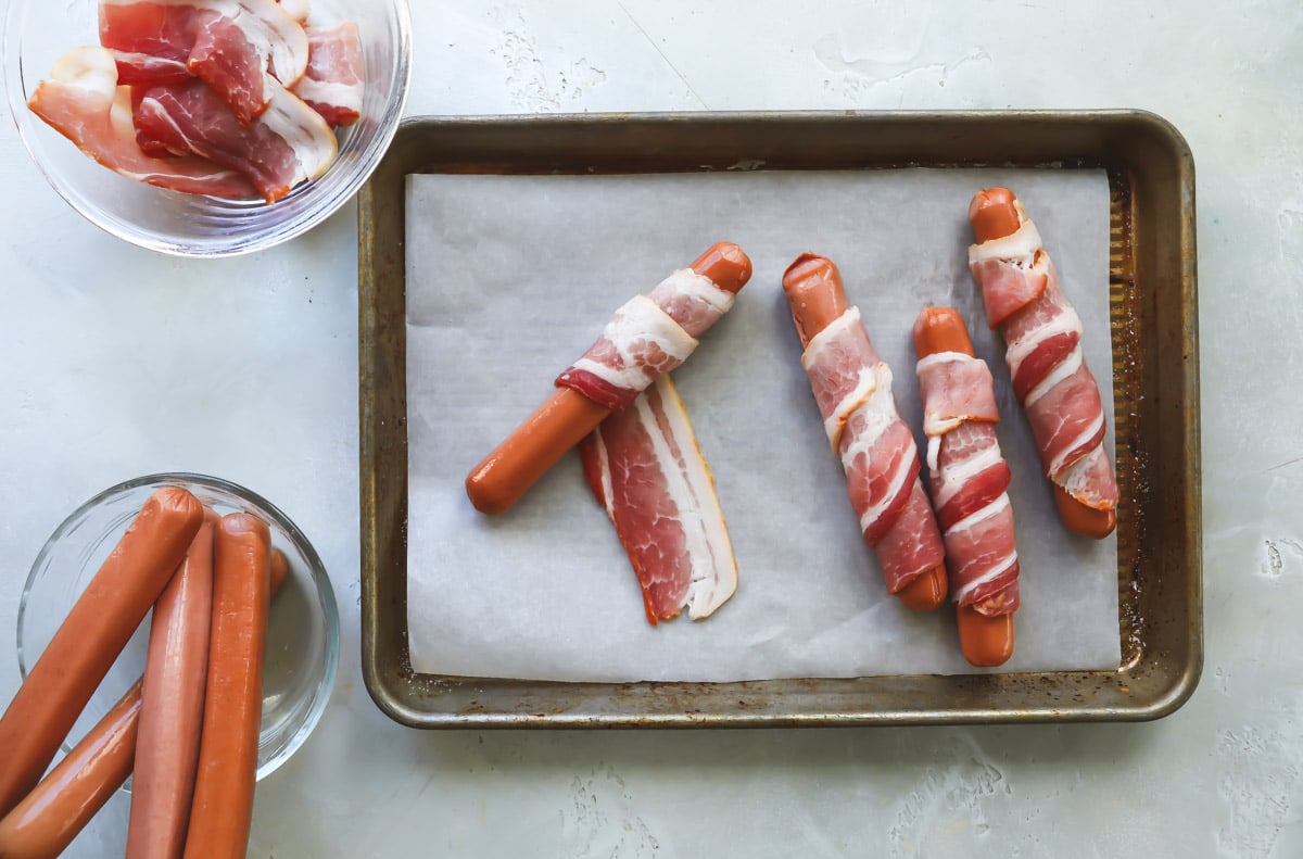 Bacon being wrapped around hot dogs on a parchment paper lined baking sheet.
