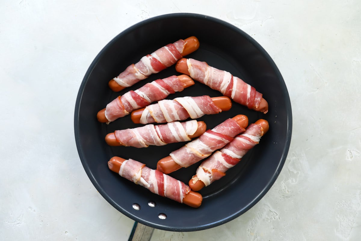 Eight bacon wrapped hot dogs in a skillet.