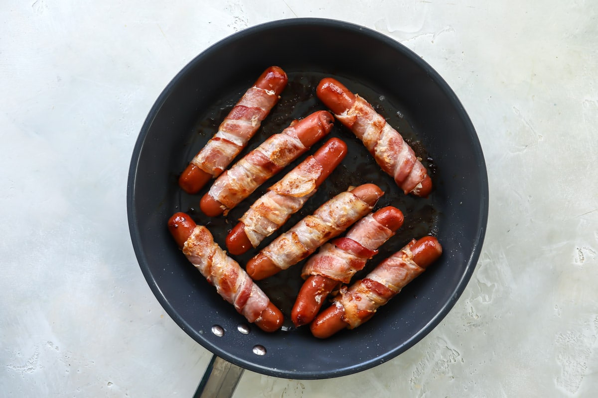 Eight bacon wrapped hot dogs in a skillet after being cooked.