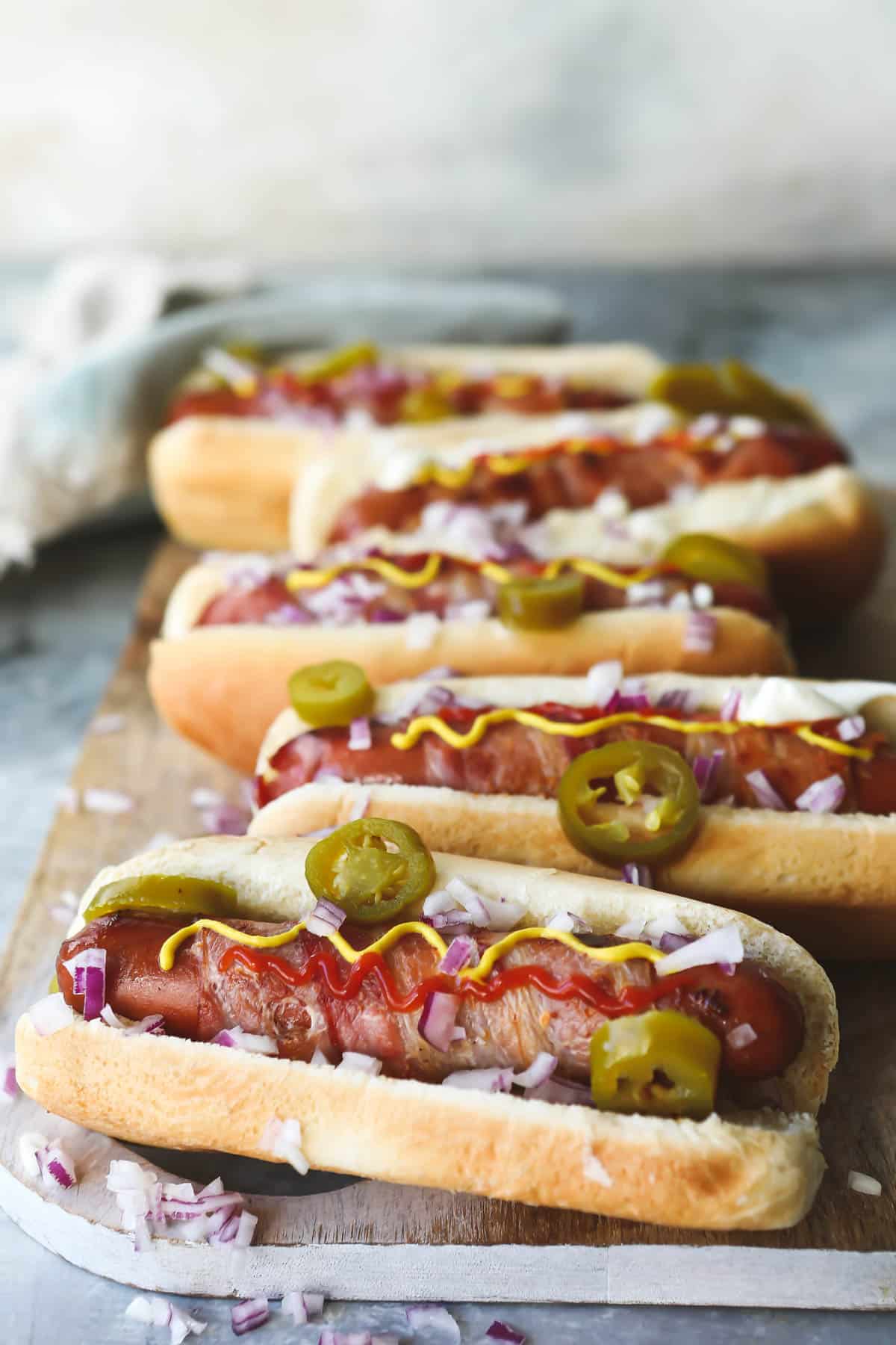 Bacon wrapped hot dogs on a wooden cutting board with toppings.