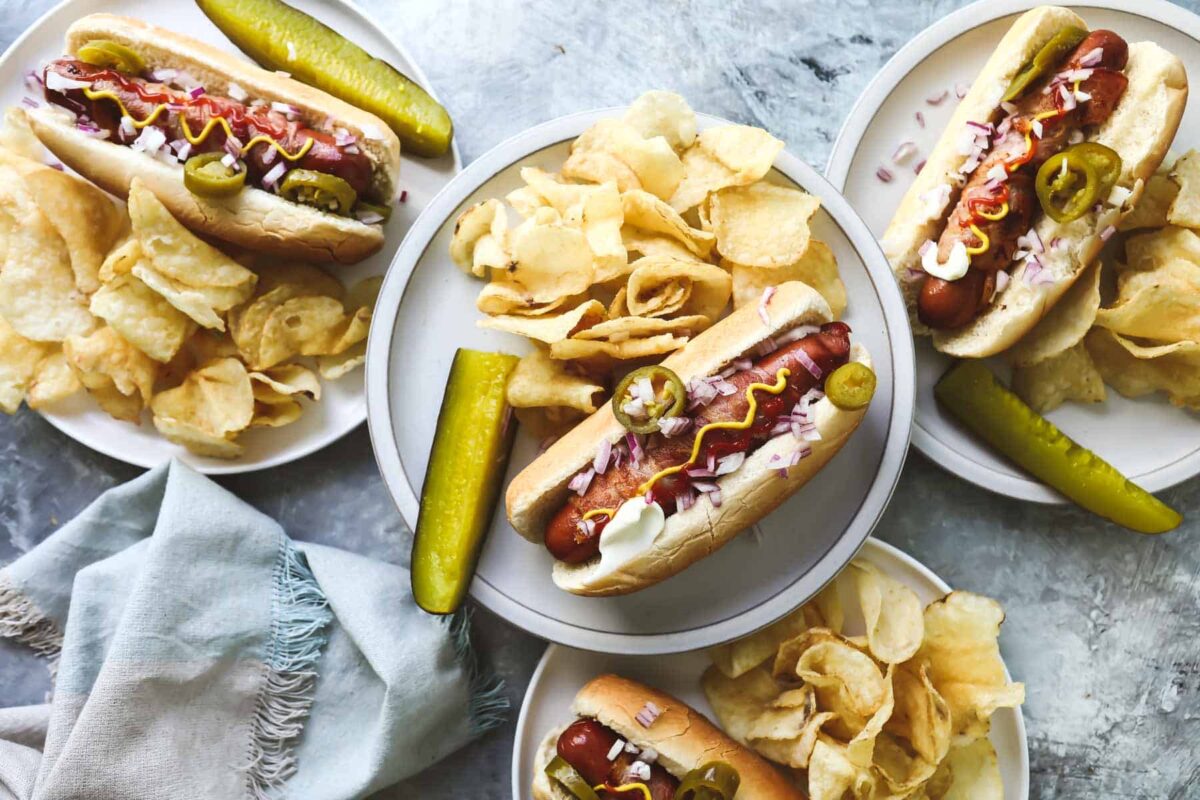 Four plates with bacon wrapped hot dogs and chips.