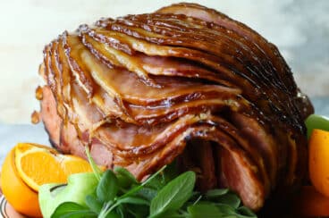 A sliced baked ham on a platter with fresh herbs and oranges.