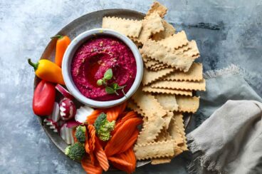 A small bowl of beet hummus surrounded by vegetables and crackers.