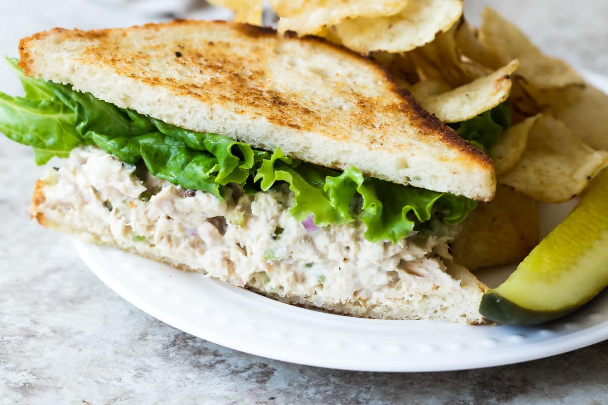 A tuna salad sandwich on a plate next to chips and a pickle spear.