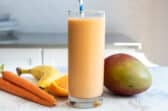 A glass of carrot smoothie next to a mango, carrots, and a banana.