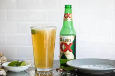 A Chelada next to a beer bottle and a small bowl of limes.