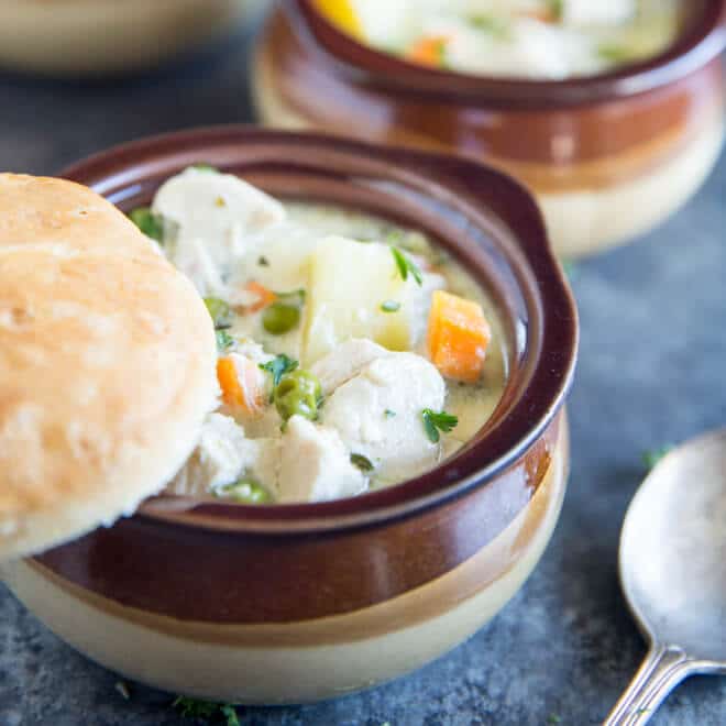 Bowl of creamy and delicious Chicken Pot Pie soup with carrot, peas, potatoes and chunks of white chicken meat in a creamy sauce with a freshly made biscut balancing on the top of the brown bowl. There is a spoon in the foreground, and a second bowl of soup in the background.