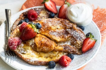 Croissant French toast on a white plate with fruit and whipped cream.