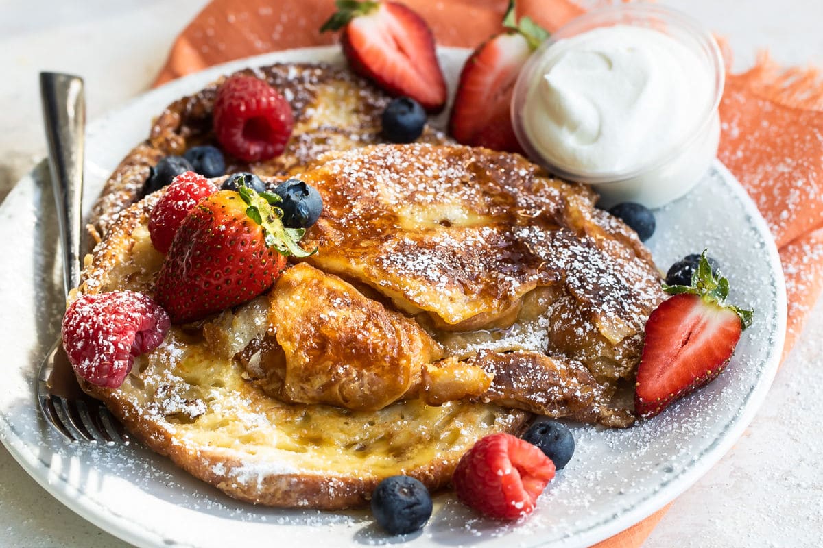 Croissant French toast on a white plate with fruit and whipped cream.