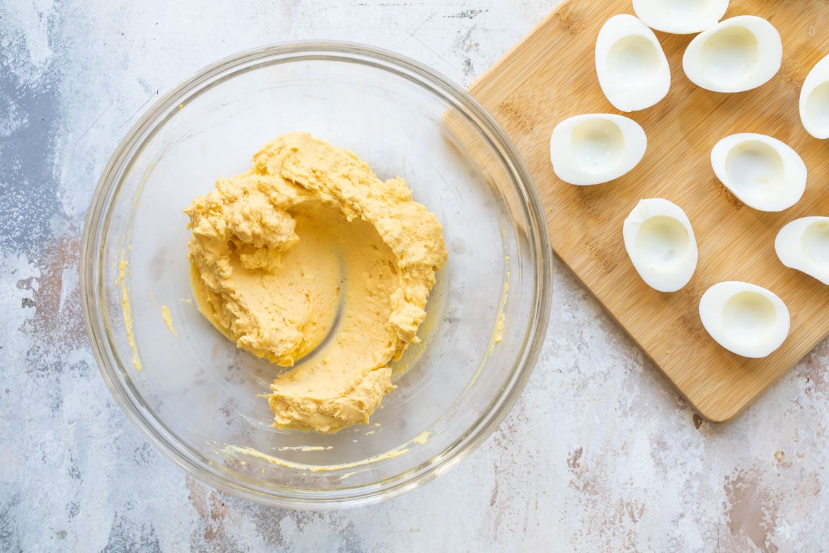Deviled egg filling in a clear bowl next to a wooden cutting board with hard boiled egg white halves on it.