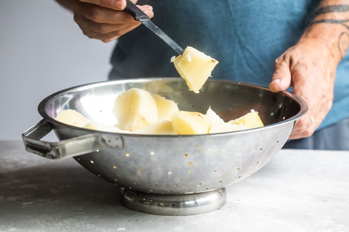 A colander with chunks of boiled potatoes (one pierced by a knife).