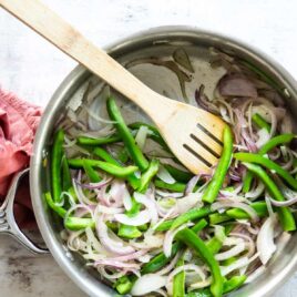 Fajita veggies cooking in a silver skillet with a wooden utensil.