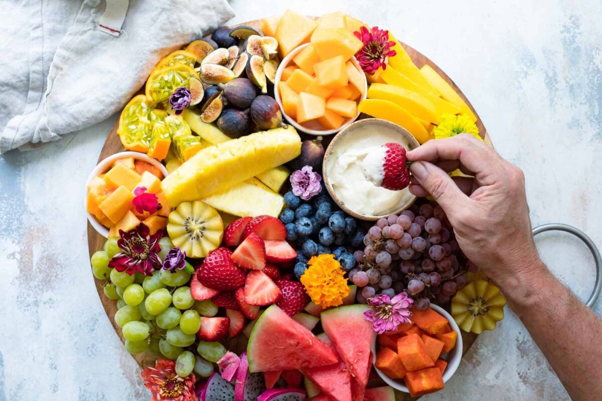 Someone dipping a fresh strawberry into a bowl of fruit dip on a fruit platter.