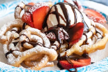 A funnel cake topped with strawberries, ice cream, and chocolate syrup on a paper plate.