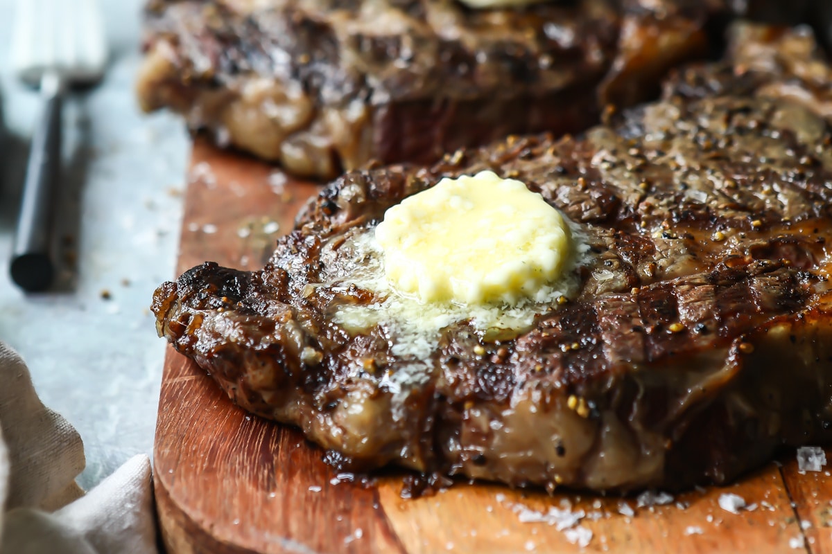 Two Rib Eye steaks on a wooden cutting board topped with butter.