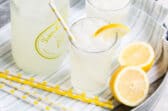 Lemonade in glasses on a silver platter with yellow and white straws and lemon garnish.