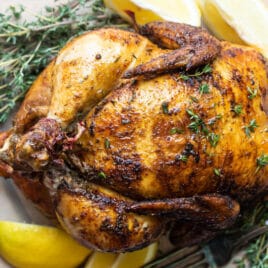 A rotisserie chicken on a white plate with slices of lemon.