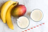 Two glasses of mango smoothie next to a mango and some bananas.