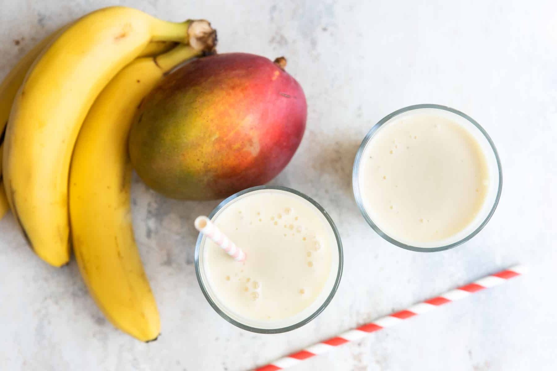 Two glasses of mango smoothie next to a mango and some bananas.