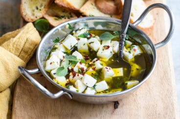 Marinated feta in a silver bowl on a wood board with slices of bread.