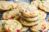 A dozen M&M cookies on a wire rack with a glass of milk and a bowl of M&Ms in the background.