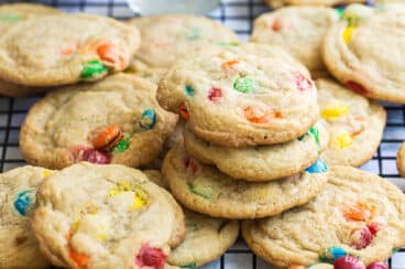 A dozen M&M cookies on a wire rack with a glass of milk and a bowl of M&Ms in the background.