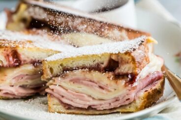 A monte cristo sandwich on a white platter being dusted with powdered sugar.