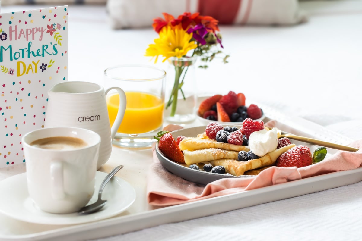 A Mother's Day breakfast tray with crepes, orange juice, coffee, fresh flowers, and a greeting card.