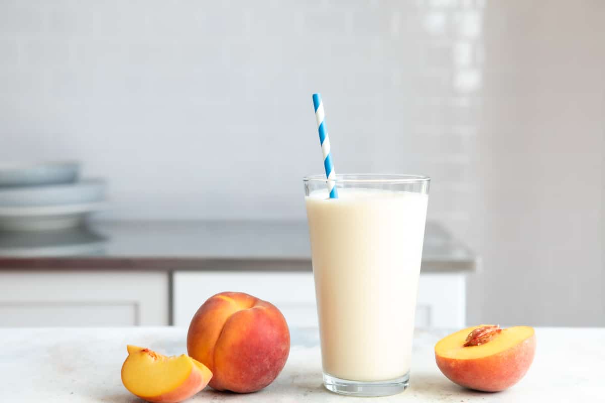 A glass of peach smoothie next to fresh peaches on a counter.