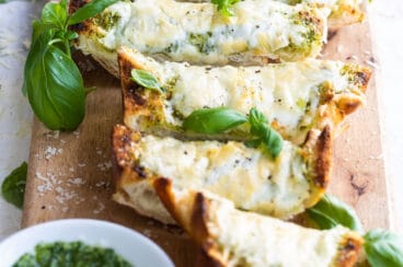Slices of pesto cheese bread surrounded by basil leaves on a wooden cutting board.