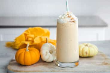 A pumpkin smoothie in a clear glass surrounded by small pumpkins.