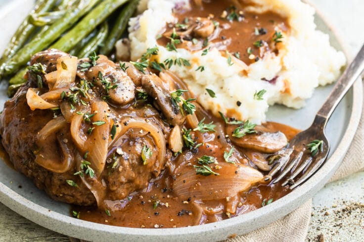A salisbury steak on a plate with mashed potatoes, gravy, and green beans.