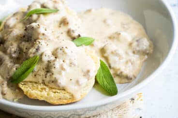 A white bowl of biscuits smothered in sausage gravy garnished with sage leaves.