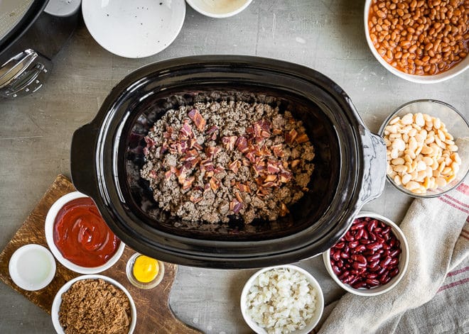Slow cooker calico beans ingredients in a black slow cooker and in various bowls.
