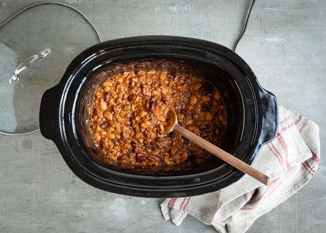Slow cooker calico beans in a black slow cooker with a wooden spoon.