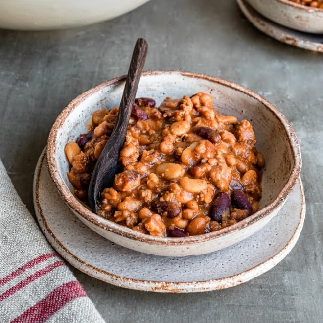 Slow cooker calico beans in a white stone bowl with wooden spoon.