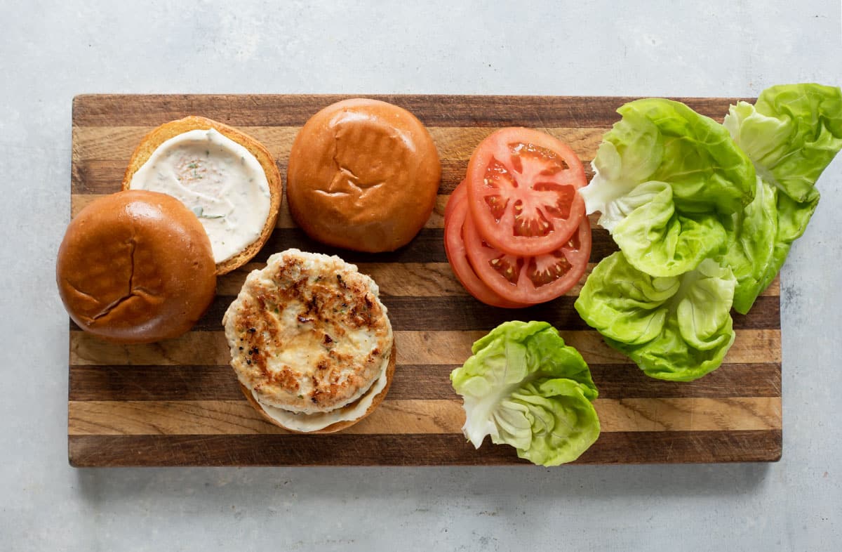 Buns, turkey burgers, lettuce and tomatoes on a wooden cutting board.