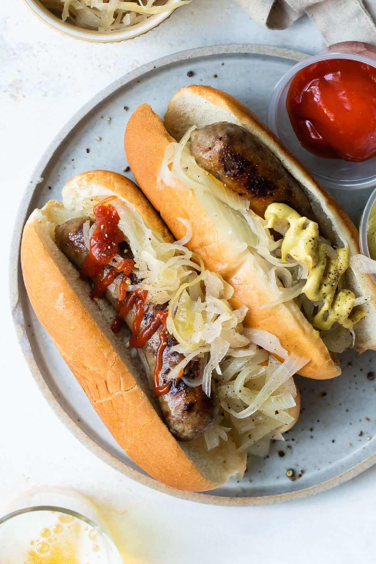 A plate of beer brats with ketchup, mustard, onions, and sauerkraut.