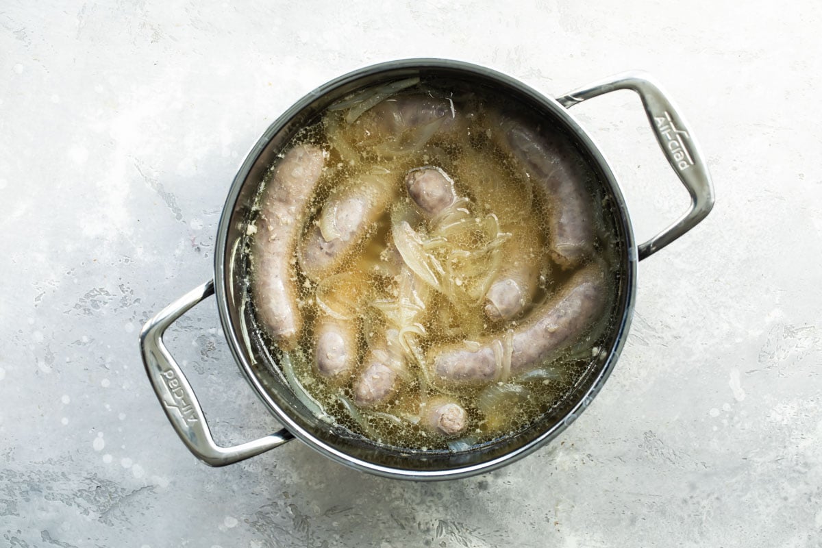 Brats boiling in beer and onions in a metal pot.