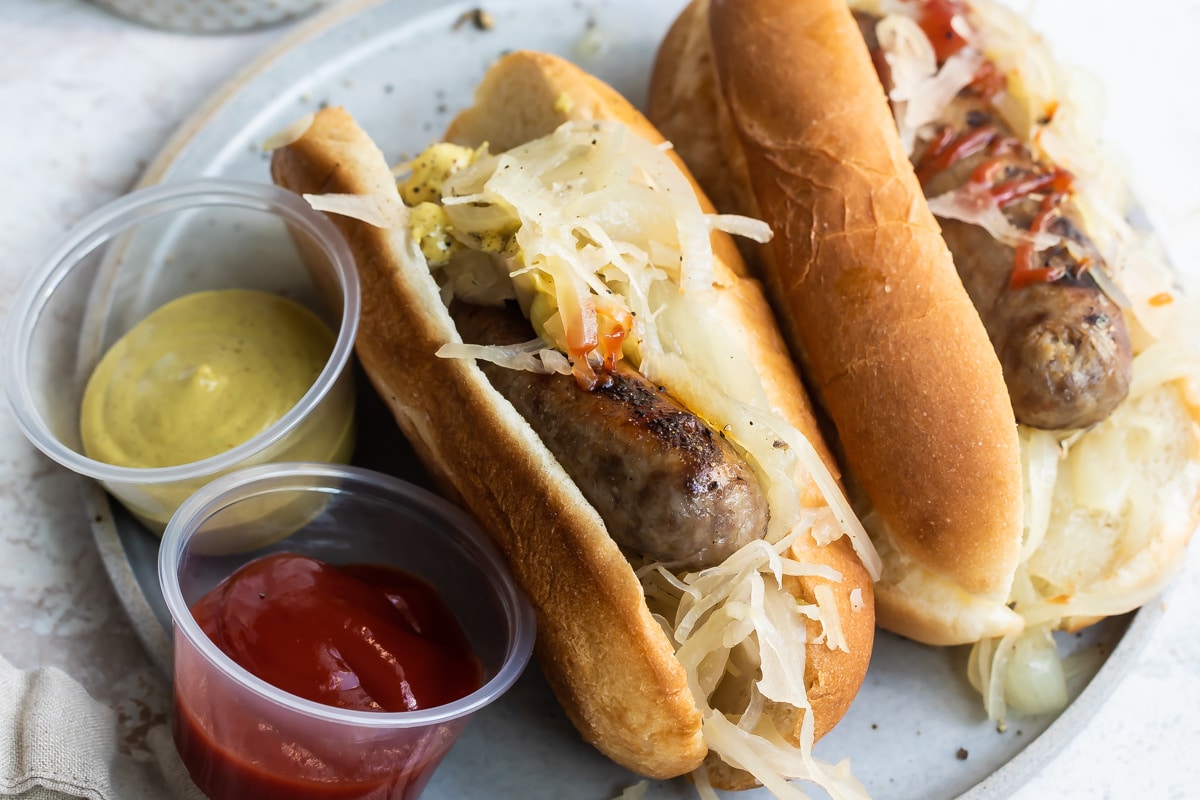 A plate of beer brats with ketchup, mustard, onions, and sauerkraut.